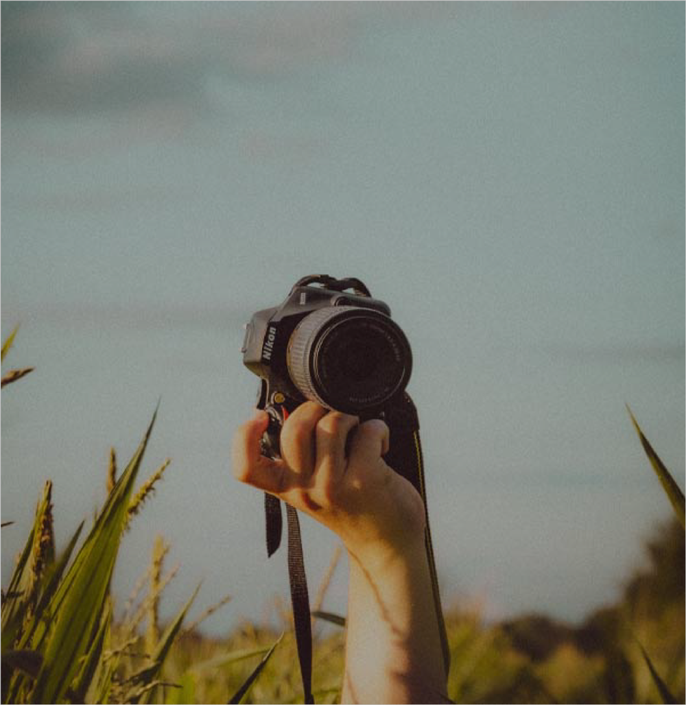 Person holding a DSLR camera above tall grass, with a blue sky in the background.