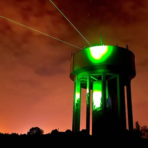 A tall structure emits green laser beams against a cloudy, orange-tinted night sky. Trees and silhouettes are visible in the background.