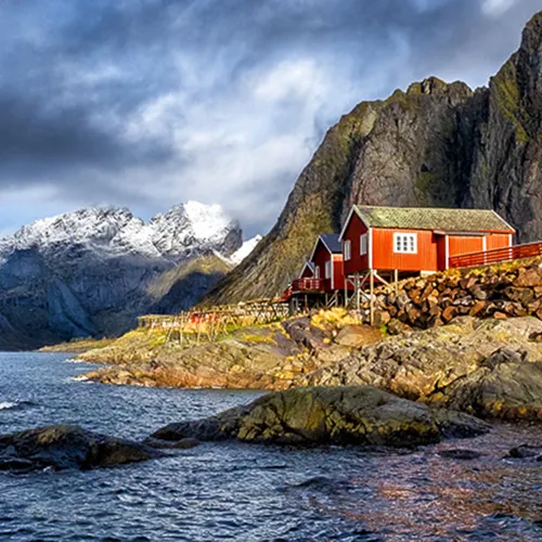 Red houses on rocky shoreline with mountain range and snow-capped peaks in the background under a cloudy sky.