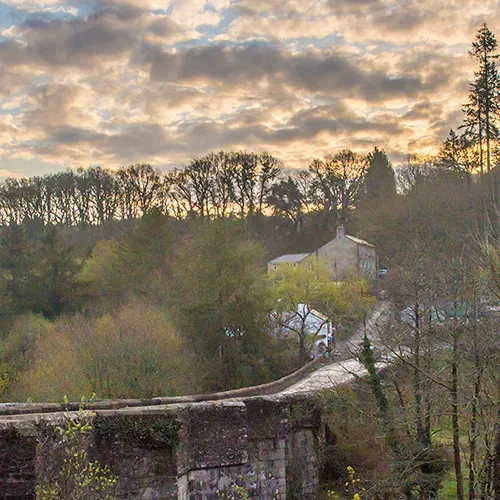 A stone bridge extends over a lush, tree-filled valley with a house in the background under a cloudy sky at sunset.