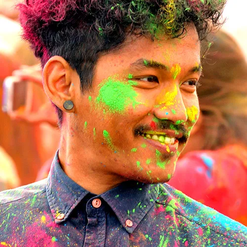 A person with a smiling face, covered in vibrant colored powders, likely participating in a Holi festival celebration.