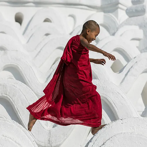 A young person in a red robe balances while walking on white architectural waves.