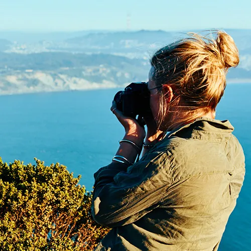 Person with blonde hair in a bun taking a photo of a scenic ocean view, with mountains in the background.