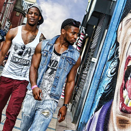 Two men walking down a street, one wearing a cap and tank top, the other in a denim vest and ripped jeans, with colorful graffiti nearby.