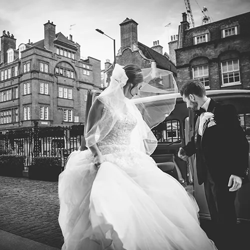 A bride and groom dressed in wedding attire stand beside a car on a cobblestone street lined with buildings.