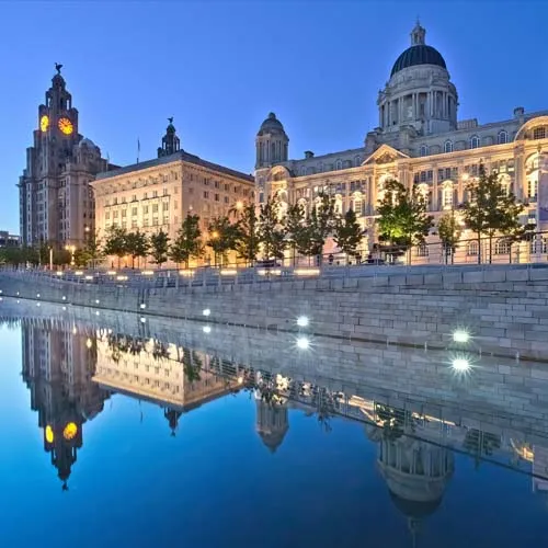 Waterfront buildings at dusk with reflections in a calm river, featuring clock tower and illuminated domed structure under a clear sky.