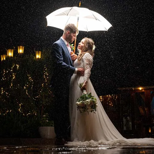 A bride and groom stand under an umbrella at night in the rain, with the bride holding a bouquet and lights in the background.