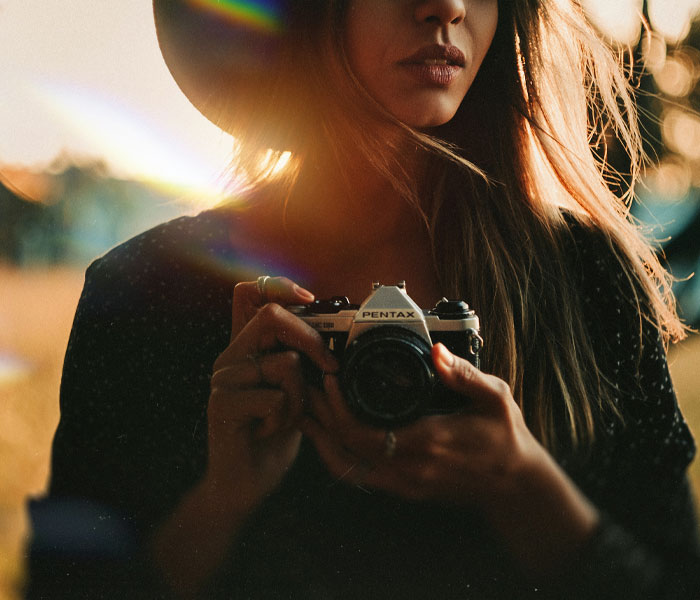 Person wearing a hat and holding a Pentax camera, with sunlight creating a lens flare effect in the background.